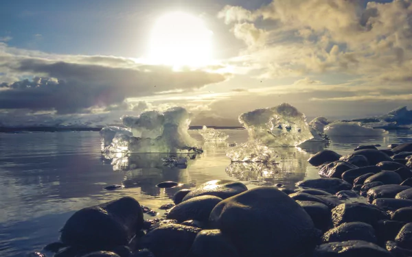 A serene Icelandic landscape featuring icebergs floating on calm water with smooth rocks in the foreground under a bright sun and cloudy sky, captured in HD.