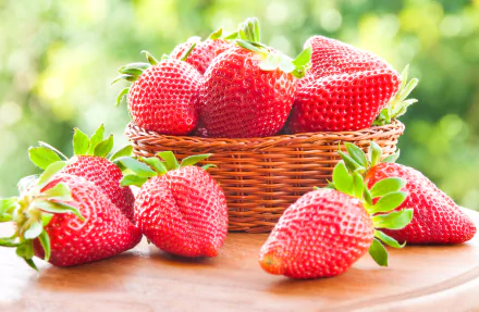 A rustic basket filled with vibrant red strawberries, surrounded by additional strawberries on a wooden surface, captured in a bright and fresh outdoor setting. 4K Ultra HD wallpaper.