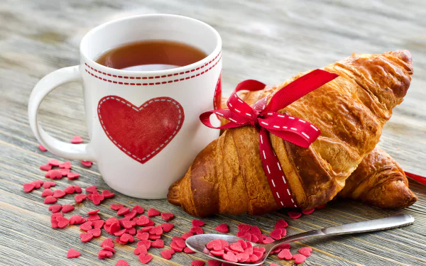 A heart-patterned mug filled with tea sits beside croissants wrapped in red ribbons, scattered with heart-shaped sprinkles, creating a warm and inviting breakfast scene.