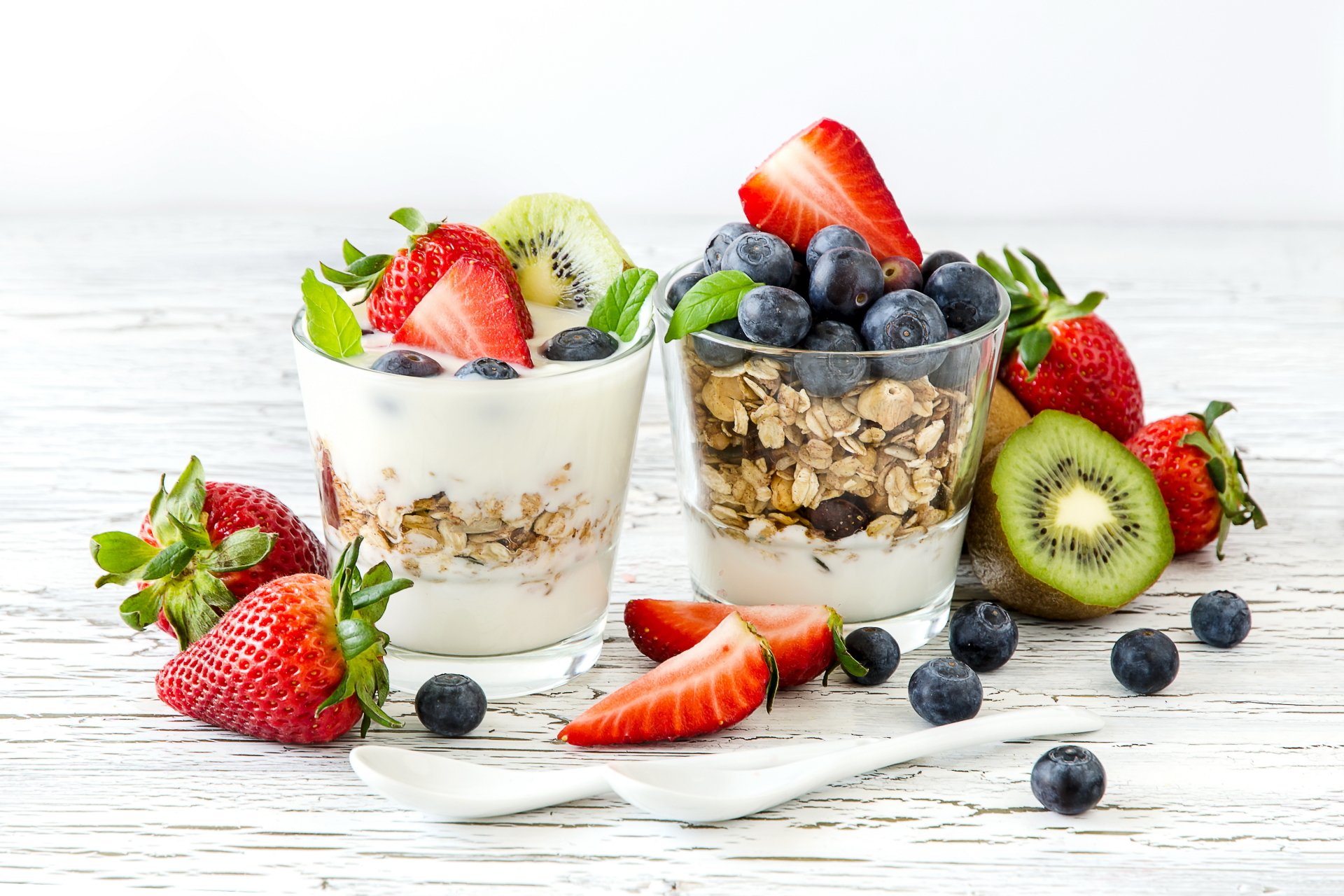 Close-up of two glasses filled with yogurt, cereal, and fresh berries including strawberries, blueberries, and kiwi, presented on a white wooden surface.