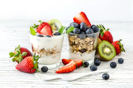 Close-up of two glasses filled with yogurt, cereal, and fresh berries including strawberries, blueberries, and kiwi, presented on a white wooden surface.
