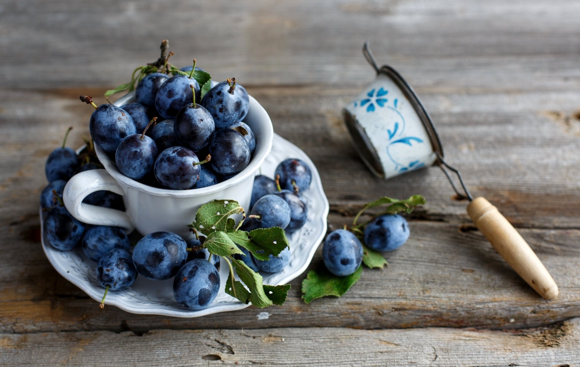 HD desktop wallpaper featuring fresh plums in a white cup and saucer, with some scattered on a rustic wooden surface alongside a vintage tea strainer.