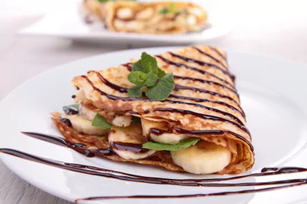 A delicious crêpe filled with banana and drizzled with chocolate, garnished with fresh mint. The backdrop enhances the tempting presentation of this mint chocolate banana treat.