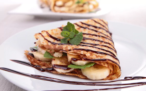 A delicious crêpe filled with banana and drizzled with chocolate, garnished with fresh mint. The backdrop enhances the tempting presentation of this mint chocolate banana treat.