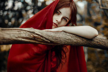 A redhead woman with striking blue eyes leans on a tree branch outdoors, wrapped in a red cloak, captured in a moody, high-resolution 4K Ultra HD wallpaper style.