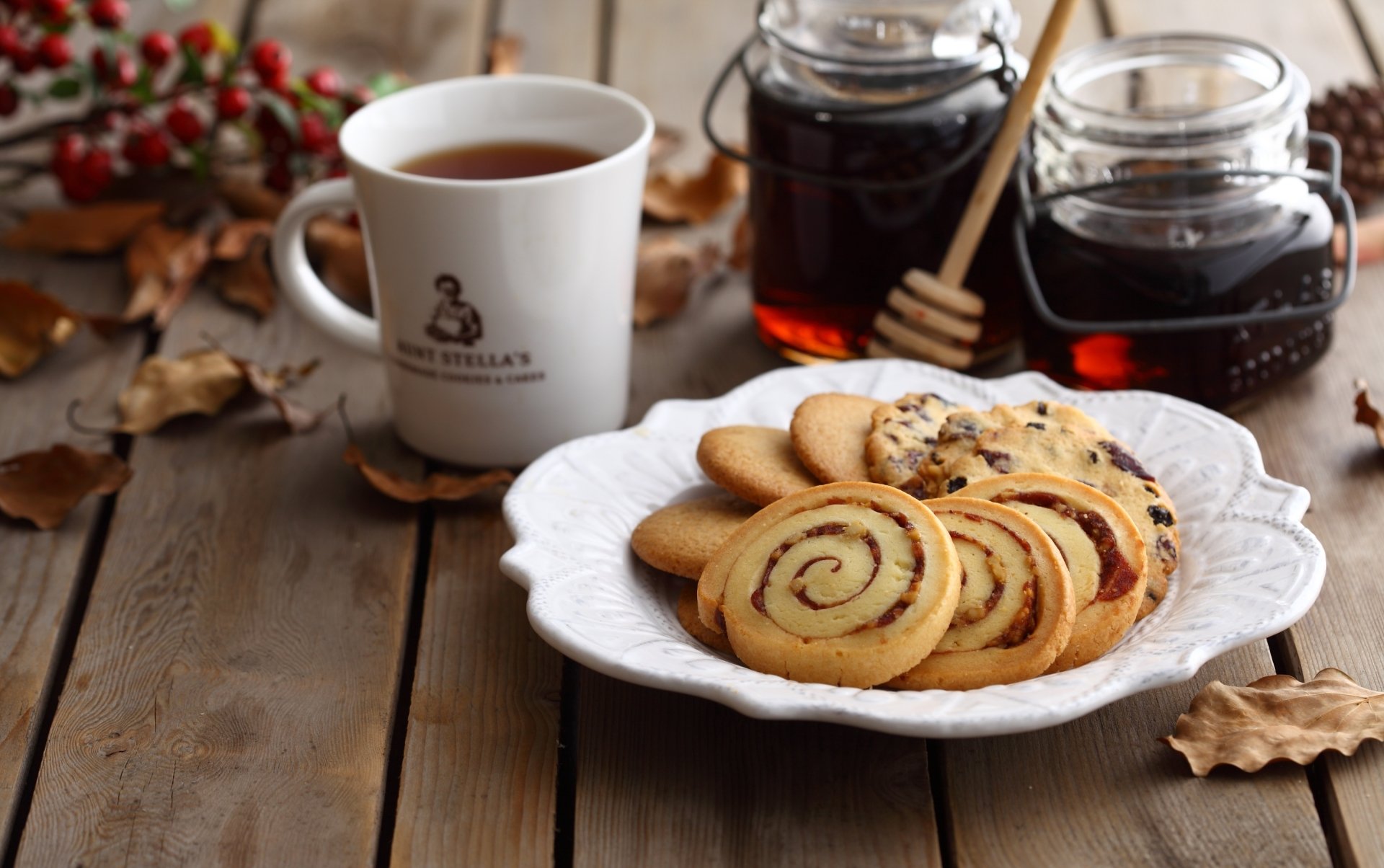 HD desktop wallpaper showing a white plate of assorted biscuits with a cup of tea and jars of honey on a wooden table surrounded by autumn leaves.