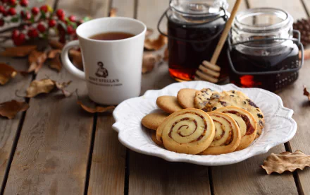 HD desktop wallpaper showing a white plate of assorted biscuits with a cup of tea and jars of honey on a wooden table surrounded by autumn leaves.