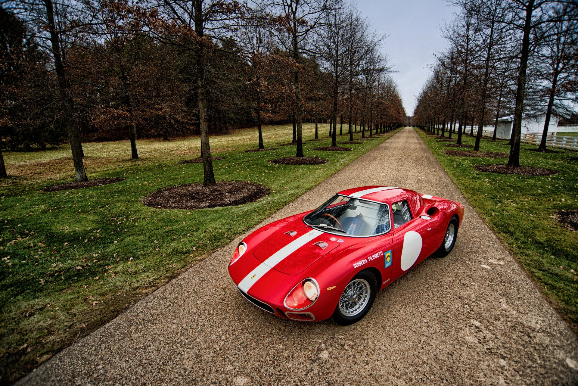 A red Ferrari 250 LM with white racing stripes parked on a tree-lined gravel road, captured in high-definition as a PC desktop wallpaper background.
