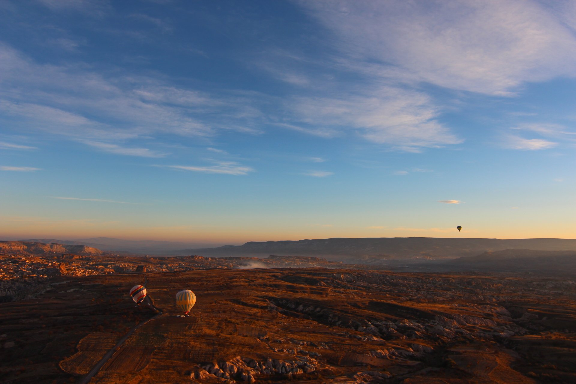 4K Ultra HD landscape of a mountainous region in Turkey with a wide sky, hot air balloons floating, and a vehicle visible on the rugged terrain at sunrise.