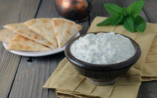 A bowl of creamy tzatziki sauce with sliced pita bread on a plate, set on a wooden table, captured in 4K Ultra HD as a PC desktop wallpaper and background.