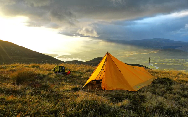 A vibrant orange tent set against a stunning mountain landscape, bathed in golden light, captures the essence of camping in nature. A beautiful 4K Ultra HD wallpaper scene.
