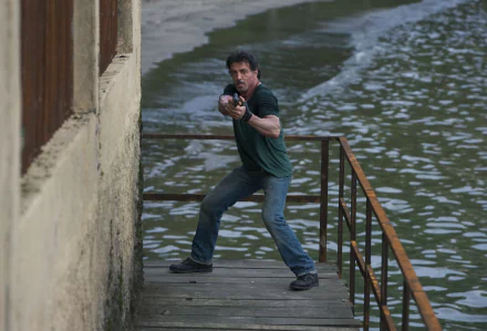 Sylvester Stallone as Barney Ross in The Expendables, poised with a gun on a dock by the water. A striking 4K Ultra HD desktop wallpaper featuring action and intensity.