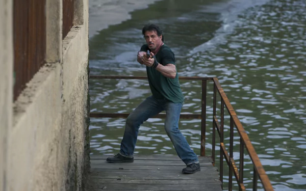 Sylvester Stallone as Barney Ross in The Expendables, poised with a gun on a dock by the water. A striking 4K Ultra HD desktop wallpaper featuring action and intensity.