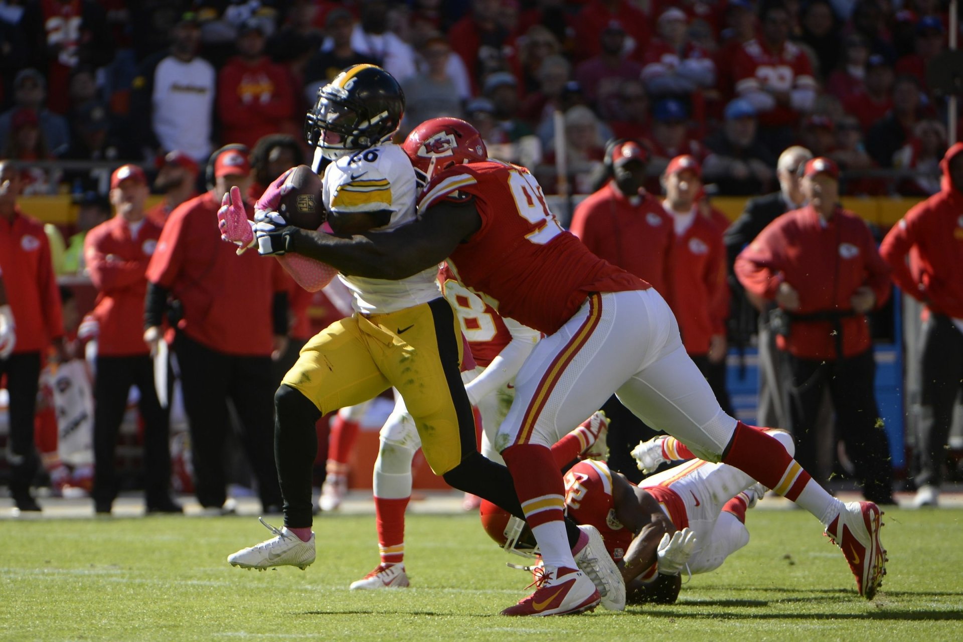 Sports HD PC desktop wallpaper background: Kansas City Chiefs defender tackles an opposing player on a sunny stadium field as teammates and fans watch from the stands.