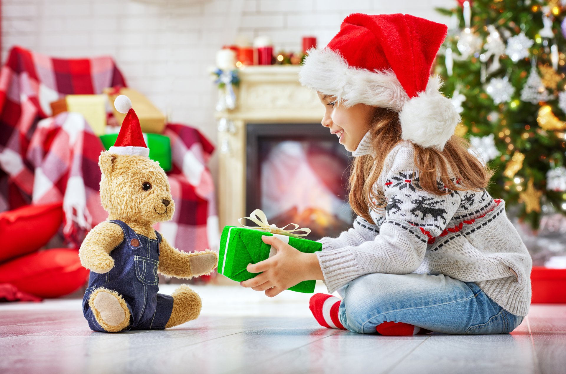 A child wearing a Santa hat gives a gift to a teddy bear also dressed in holiday attire, captured in a festive 4K Ultra HD Christmas desktop wallpaper scene.