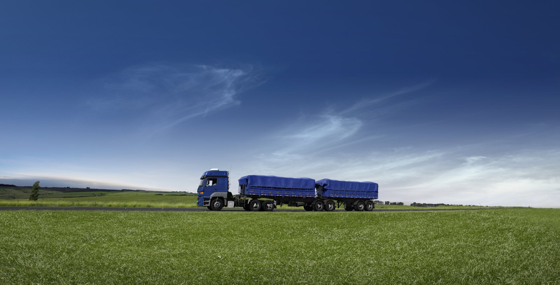 8K Ultra HD image of a blue truck with a double trailer driving across a green field under a clear blue sky, featured as a PC desktop wallpaper.