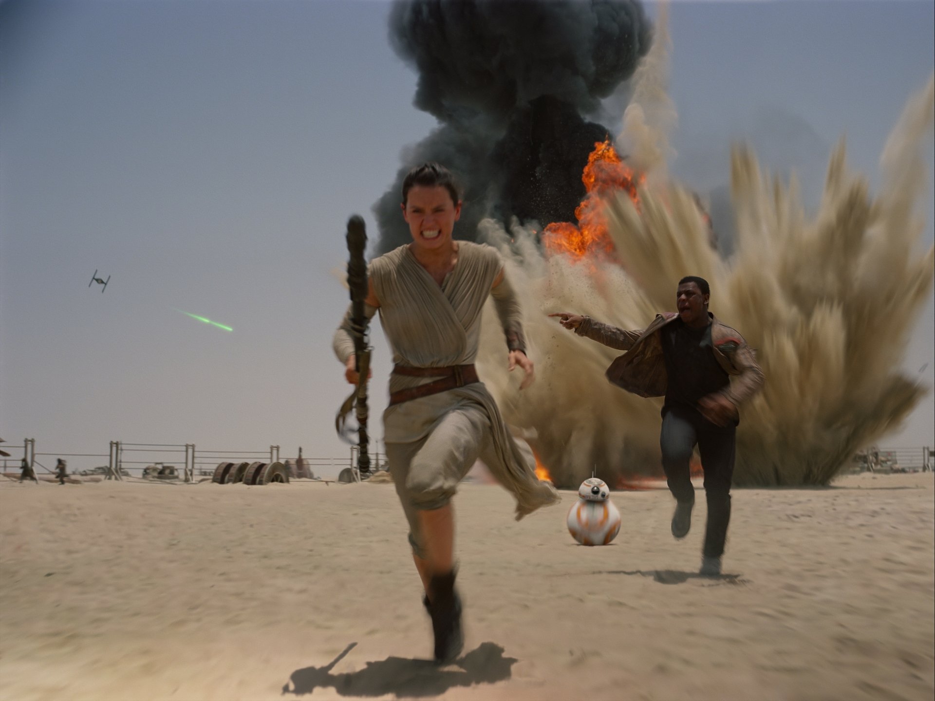 Rey and Finn from Star Wars Episode VII: The Force Awakens run through a desert amidst explosions, with BB-8 trailing behind, capturing an intense moment from the film.