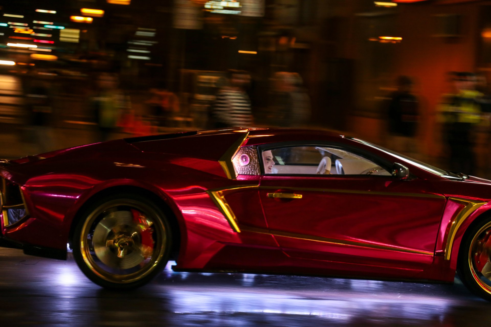 A sleek, red sports car from the movie Suicide Squad speeds through a bustling city at night, with vibrant lights reflecting off its shiny surface. HD wallpaper background.