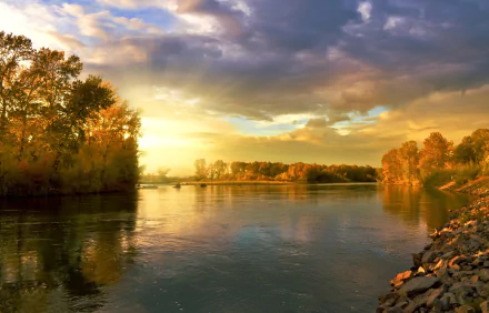 HD PC wallpaper: autumn river at sunset with golden sunbeams breaking through clouds, trees in fall colors along the water and a rocky shoreline reflecting the sky.