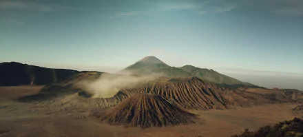 2K Quad HD desktop wallpaper of Mount Bromo, Indonesia: rugged volcanic caldera and ridged slopes beneath a pale sky with drifting mist.