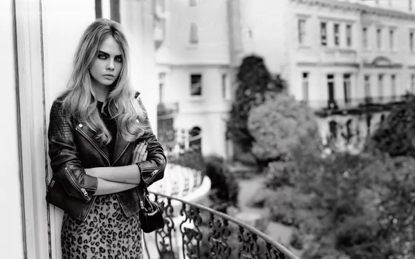 A striking black-and-white image of Cara Delevingne, leaning against a balcony railing, dressed in a leather jacket and leopard print skirt, set against a stylish urban backdrop.