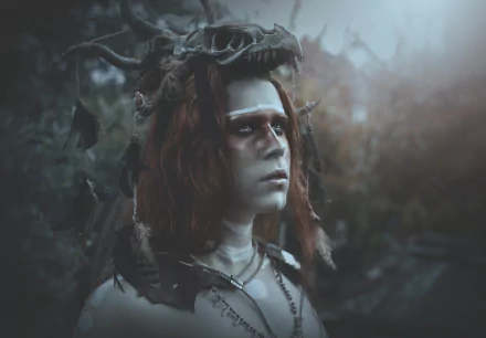 Dark artistic cosplay shaman: redhead woman model in a horned headdress and tribal paint, moody bokeh forest backdrop — HD PC desktop wallpaper/background.