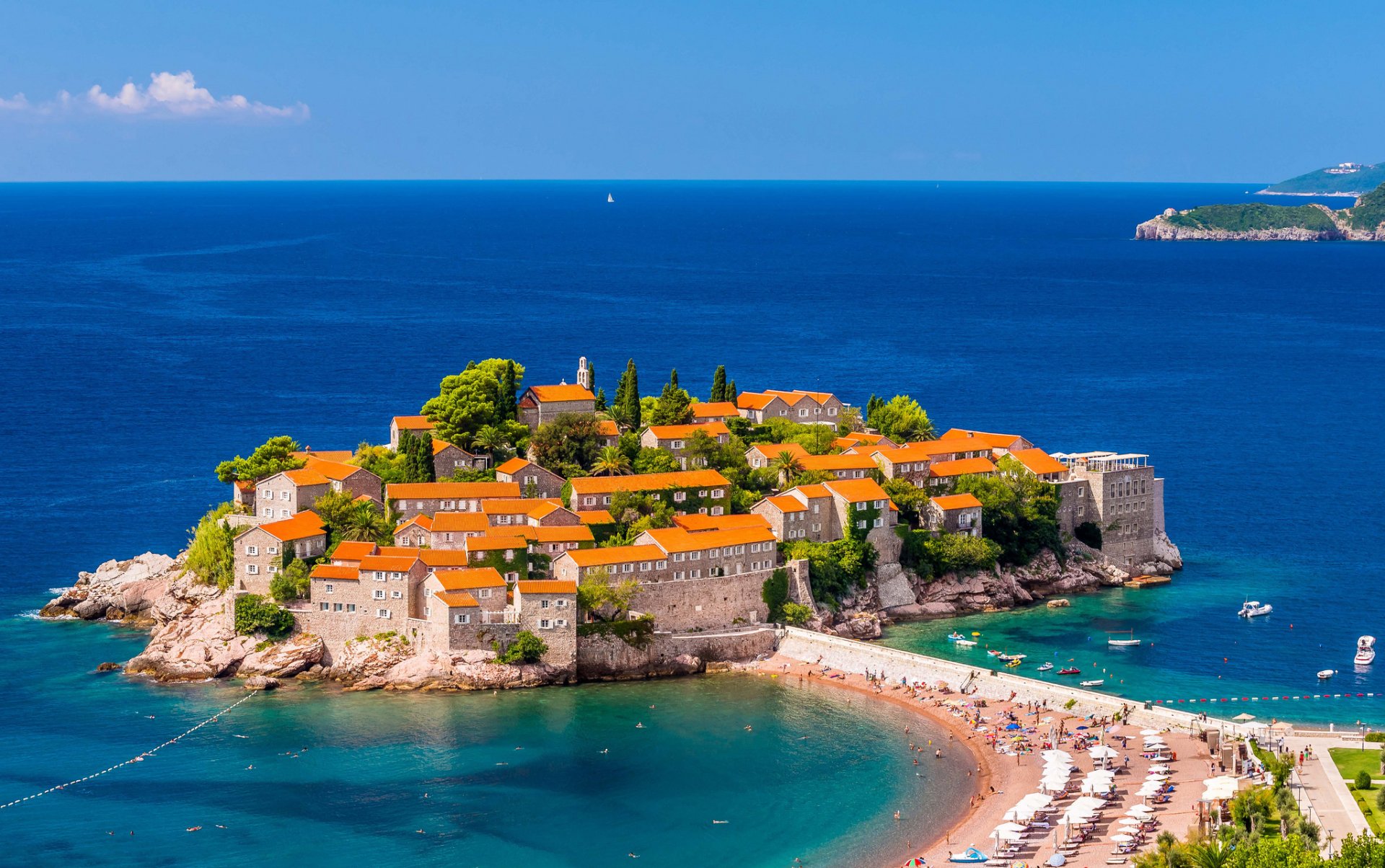 A man-made island village with orange-roofed houses on the coast of Montenegro, surrounded by blue sea and a sandy beach under a clear horizon.