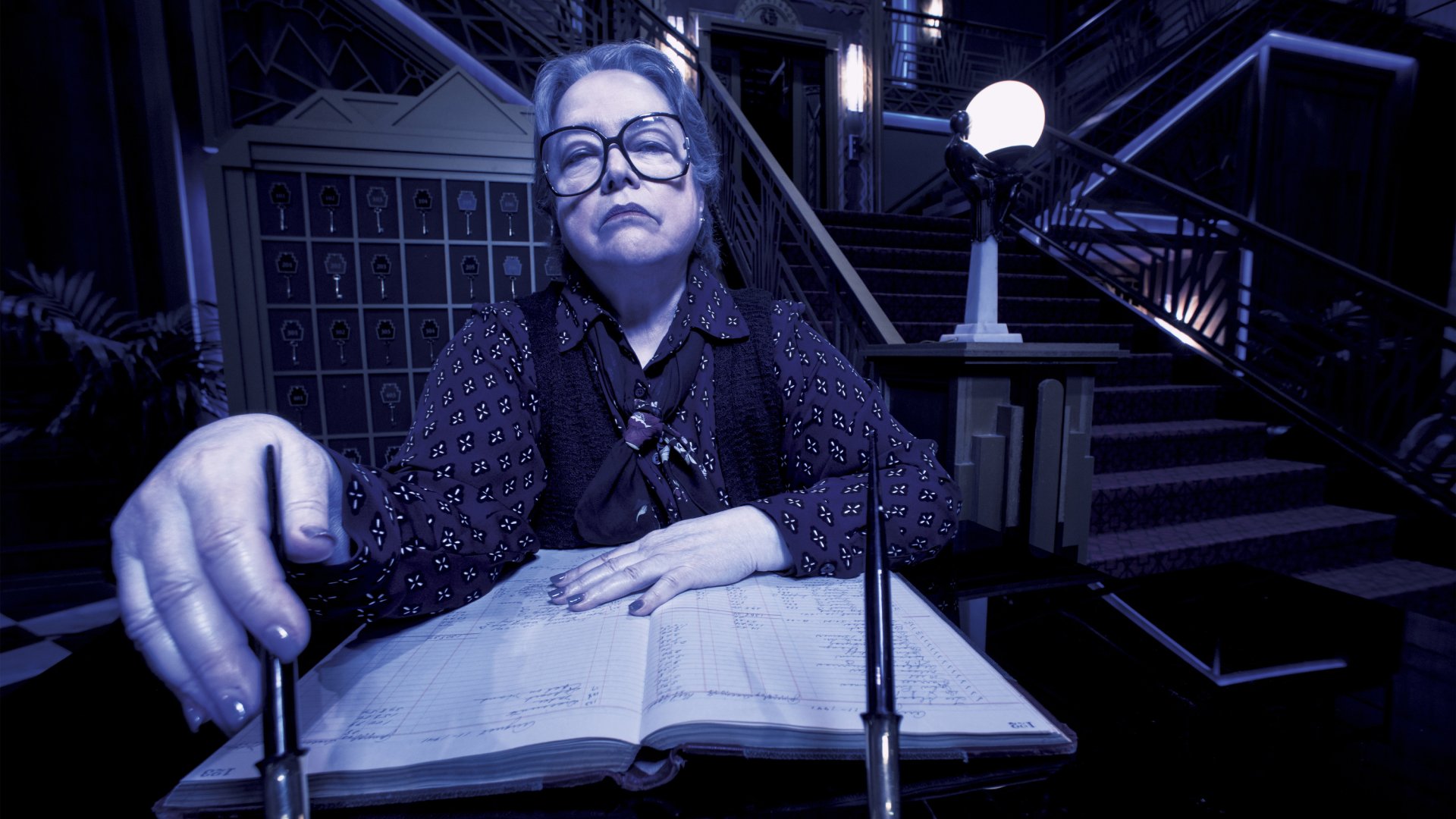 American Horror Story TV show — 2K Quad HD PC desktop wallpaper/background: blue-tinted image of a bespectacled woman leaning over an open ledger in a dim, eerie staircase hall.