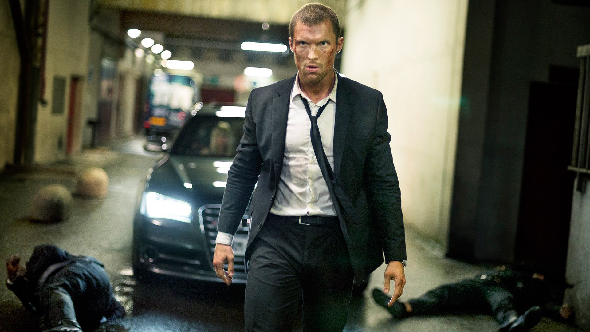 HD PC desktop wallpaper/background: action still from the movie The Transporter Refueled showing a suited man walking away from a black car in an underground garage, tense cinematic lighting.