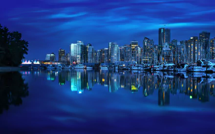 A stunning nighttime view of Vancouver's harbor, featuring a skyline illuminated in blue, with reflections shimmering on the water and yachts docked along the waterfront.