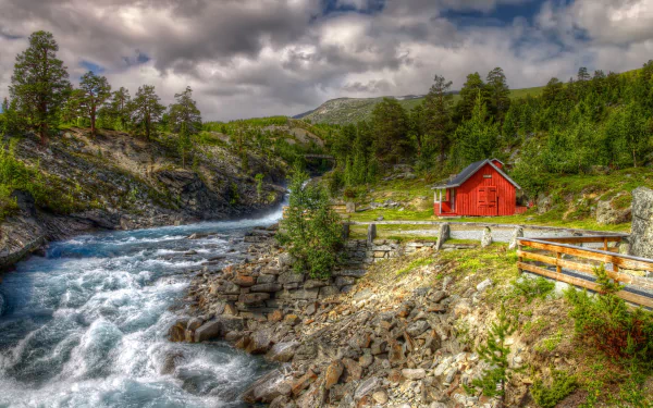 HD desktop wallpaper of a vibrant Norwegian landscape featuring a river flowing beside a red house, surrounded by trees and rocky terrain under a cloudy sky.