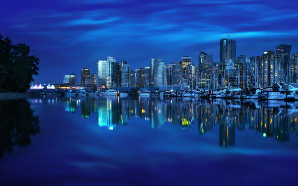 A stunning nighttime view of Vancouver's harbor, featuring a skyline illuminated in blue, with reflections shimmering on the water and yachts docked along the waterfront.