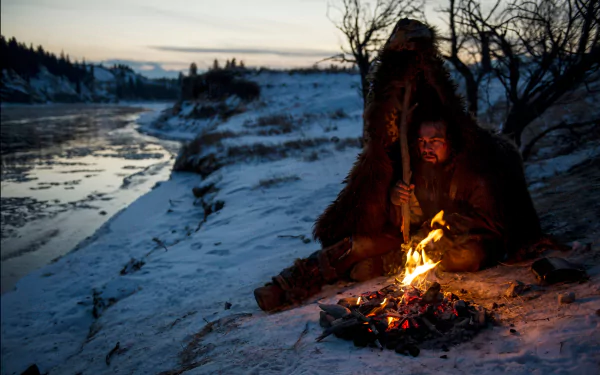 A captivating HD wallpaper featuring Leonardo DiCaprio in a scene from the movie The Revenant, depicting a rugged winter landscape with a solitary figure by a fire.