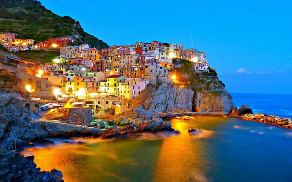 A beautiful HD wallpaper of Manarola, showcasing colorful buildings illuminated at dusk along the rocky coastline, with gentle waves reflecting warm lights against a serene backdrop.