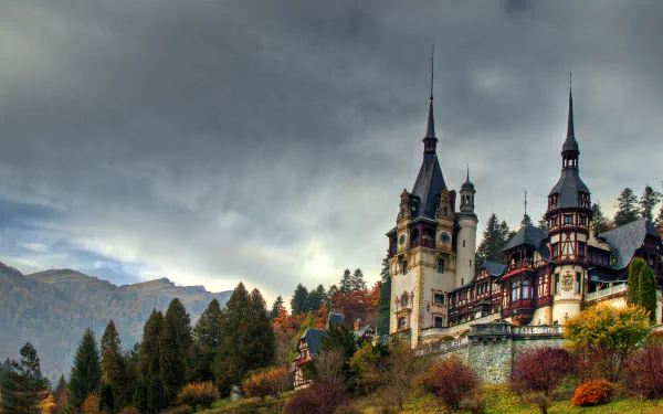 Peles Castle, a stunning man-made architectural landmark in Romania, set against a dramatic fall landscape with dense forest and cloudy skies.