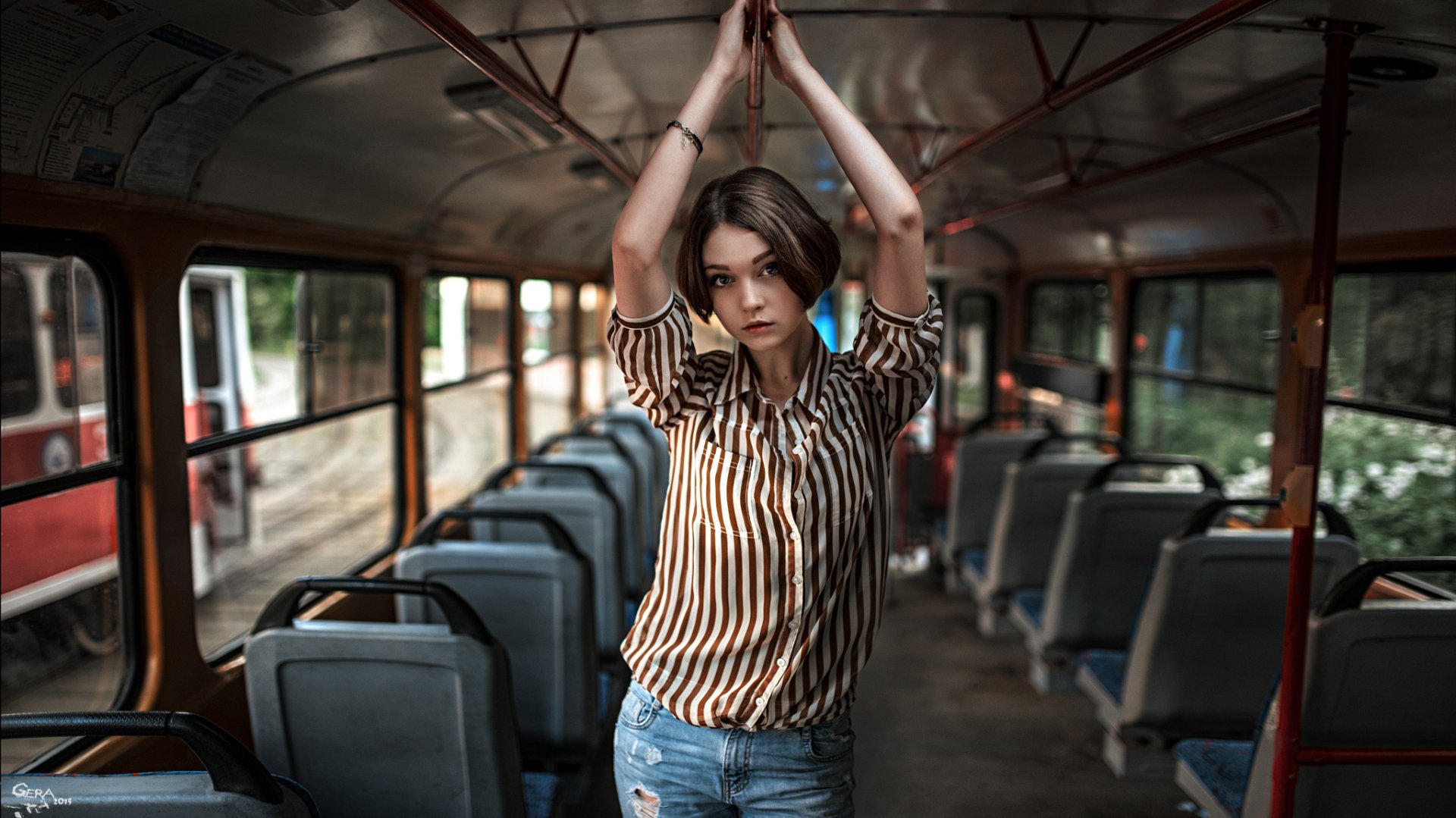A brunette woman with blue eyes poses in a bus, holding onto the overhead rail. The image features a captivating bokeh effect, creating a striking HD desktop wallpaper.