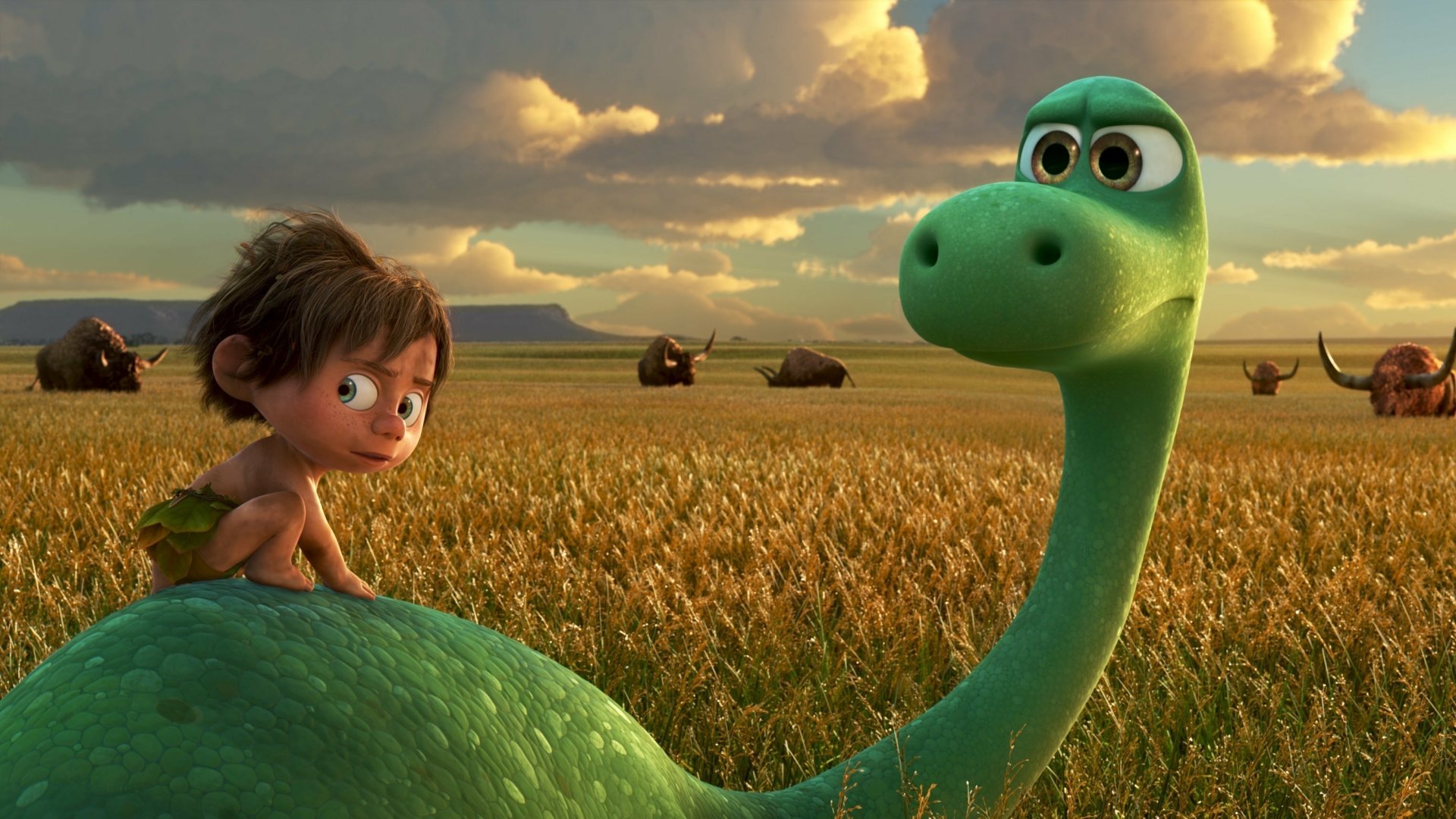 HD wallpaper from the movie The Good Dinosaur featuring a young boy and a green dinosaur in a field with cloudy skies and mountains in the background.