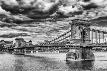 Black and white HD image of Budapest’s Chain Bridge spanning the river under a dramatic cloudy sky, showcasing the iconic man-made structure in Hungary.