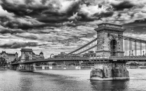 Black and white HD image of Budapest’s Chain Bridge spanning the river under a dramatic cloudy sky, showcasing the iconic man-made structure in Hungary.