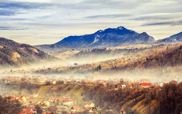 A scenic HD wallpaper of a Romanian village nestled in foggy mountain landscape, showcasing autumn colors and mist weaving through the hills.