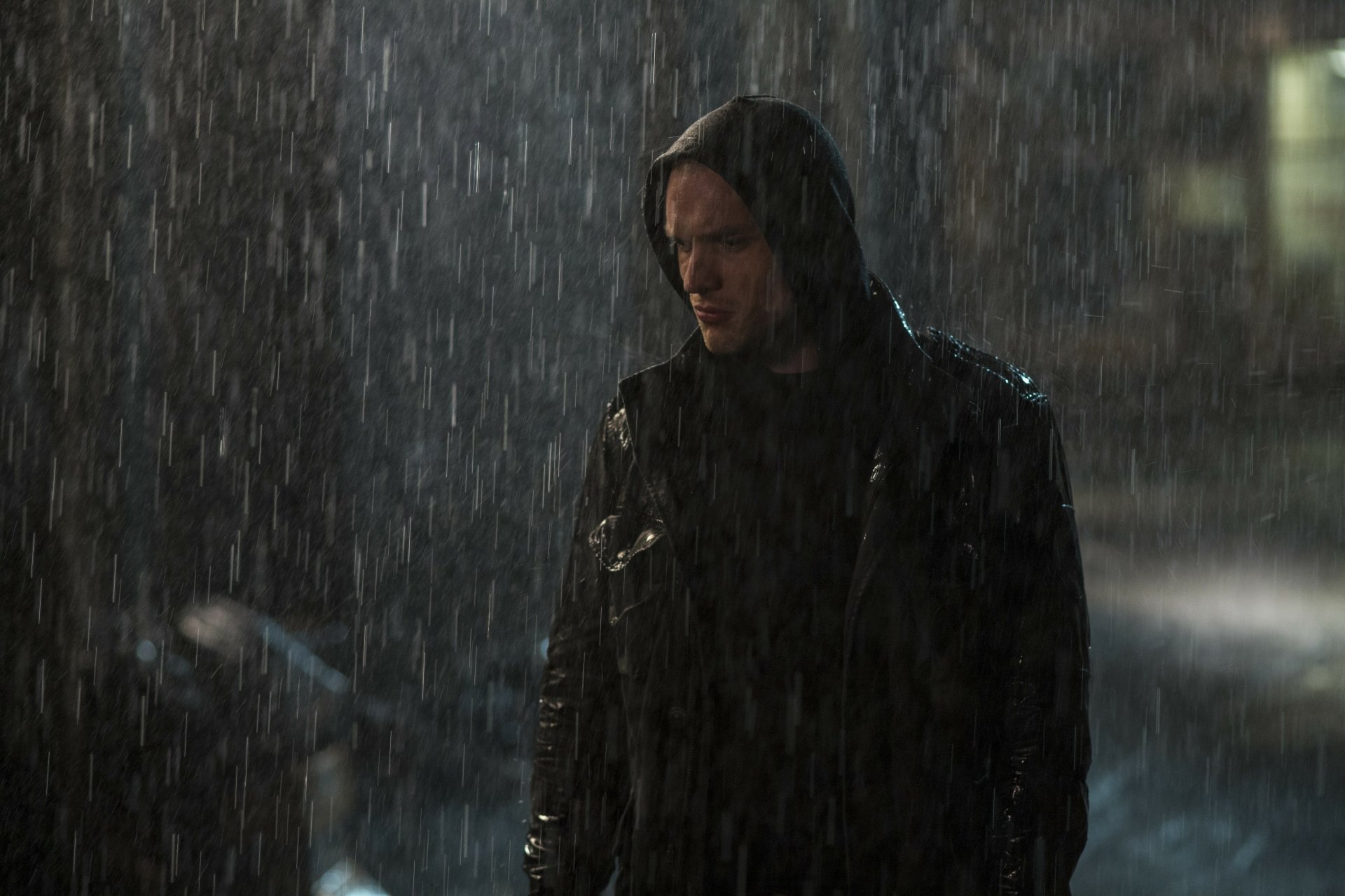 A moody scene featuring Ed Skrein in a hooded jacket, standing in heavy rain, capturing a dramatic moment from the movie Deadpool in a 4K Ultra HD background.