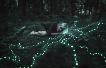 A white-haired figure lies in a dark forest surrounded by glowing blue-green bokeh lights, creating a moody, surreal atmosphere in this HD desktop wallpaper.