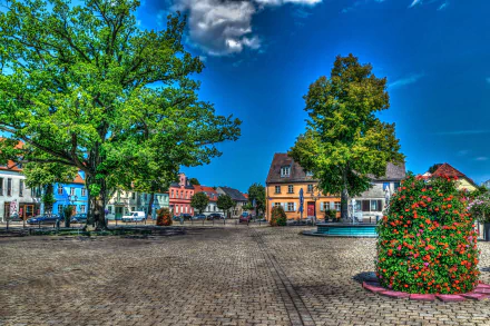 A vibrant HDR 4K Ultra HD image of a square in Brandenburg, Germany, featuring trees, colorful houses, and a clear blue sky along a peaceful town street.