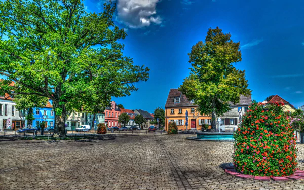 A vibrant HDR 4K Ultra HD image of a square in Brandenburg, Germany, featuring trees, colorful houses, and a clear blue sky along a peaceful town street.