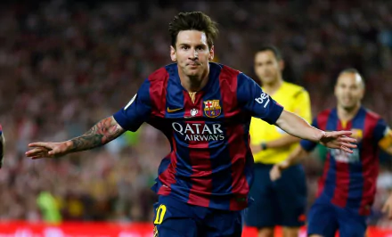 Lionel Messi wearing his FC Barcelona jersey, celebrating a goal with arms outstretched. He has a visible tattoo on his right arm. The background shows a stadium and other players.