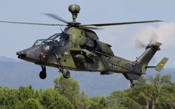 4K Ultra HD image of a military Eurocopter Tiger attack helicopter flying above green treetops with mountains in the background.