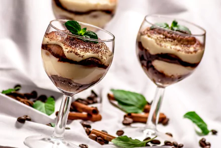 Two elegant glass cups filled with layered dessert featuring coffee beans, cinnamon sticks, and chocolate, garnished with fresh mint leaves on a bright background.