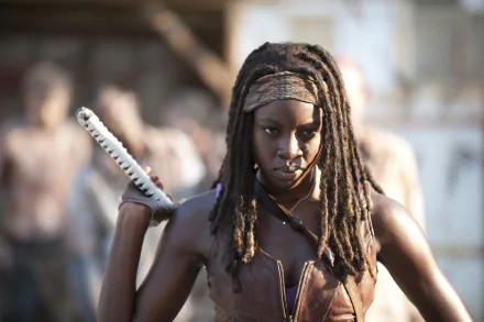 Michonne from The Walking Dead, portrayed by Danai Gurira, stands confidently with her katana, surrounded by the eerie backdrop of walkers, showcasing her strength and resolve.