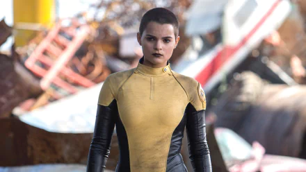 Brianna Hildebrand as Negasonic Teenage Warhead from the movie Deadpool stands confidently in front of a chaotic backdrop, showcasing her distinctive yellow and black suit.