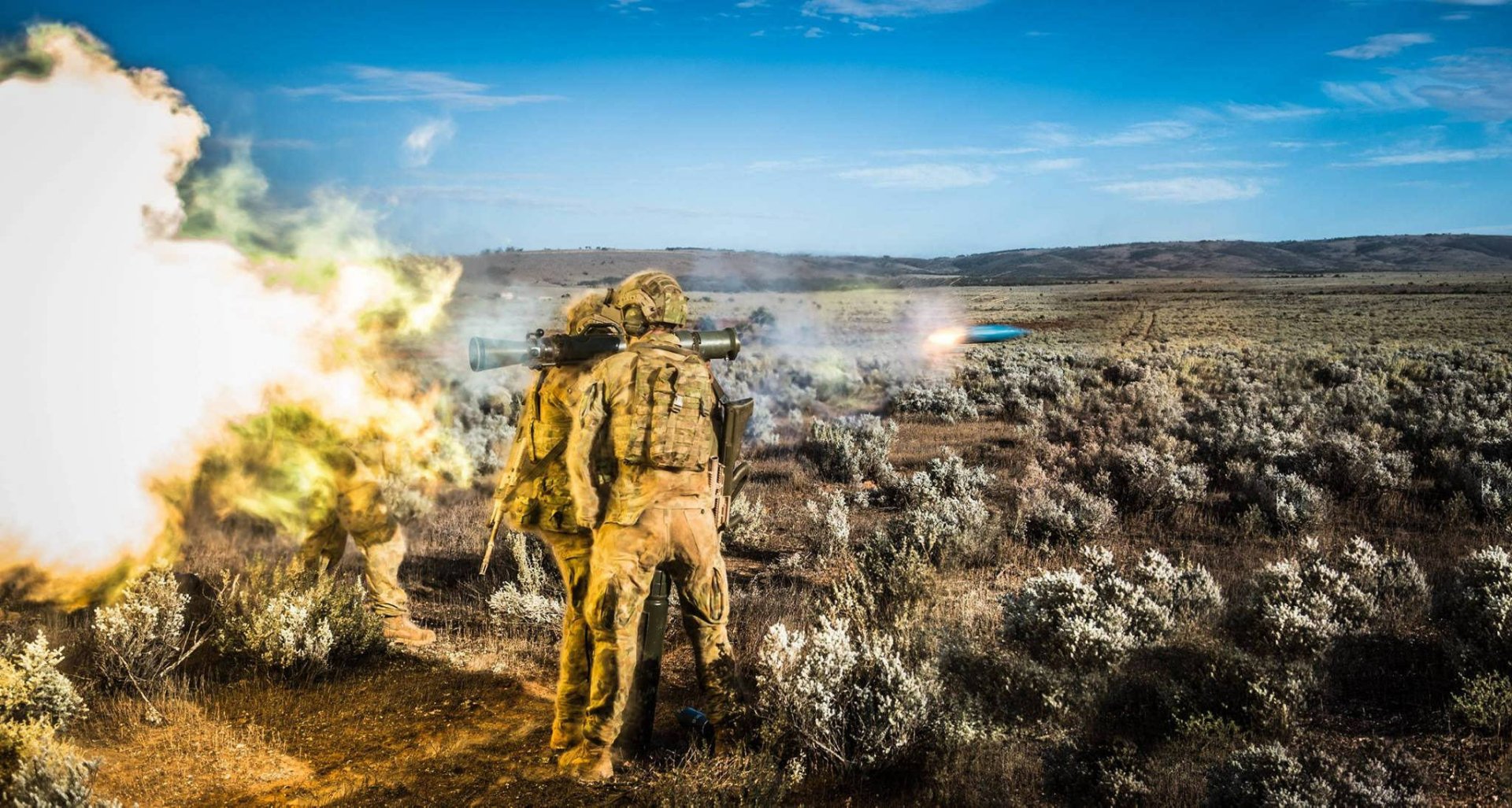 HD PC desktop wallpaper: military soldier launching a missile weapon across open scrubland, smoke and muzzle flash under a wide blue sky.